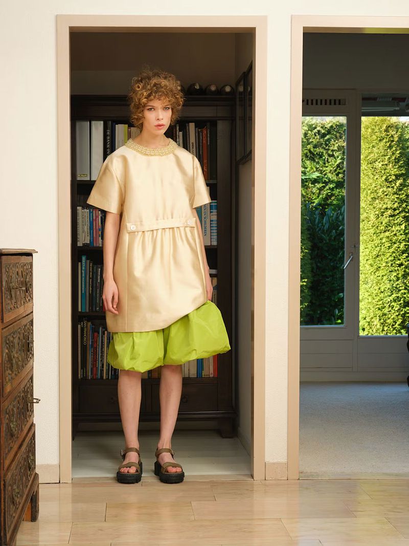 Model Bianca Huisman stands in a doorway wearing a cream satin tunic top, voluminous lime green shorts, and chunky platform sandals.