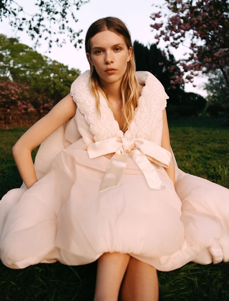 A fair-skinned young adult woman with long blonde hair wearing a light pink dress with a large bow sits in a lush green garden.