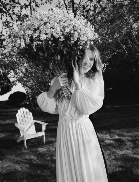 Monochrome portrait of a woman holding a large bouquet of white flowers in a garden setting, with an Adirondack chair nearby.