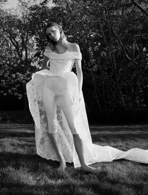 A woman poses in a white lace off-shoulder top, capri pants, and train in a lush outdoor setting with dense trees in black and white.