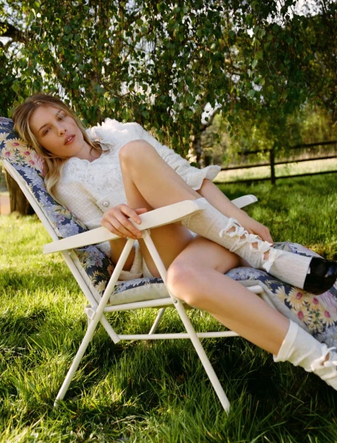 Young woman lounges in a floral deckchair outside on a sunny day with a tree in the background, wearing white lace socks and shoes.