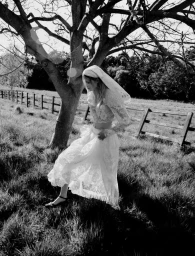 Model in a lace wedding dress and veil walking through a grassy field next to a tree, shot in monochrome.