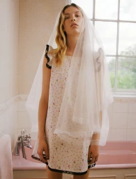 Model wearing a sheer white veil and patterned dress standing in a bathroom with a pink bathtub