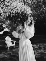 Model holding a large bouquet of white flowers in a white dress outdoors with a wooden chair in the background