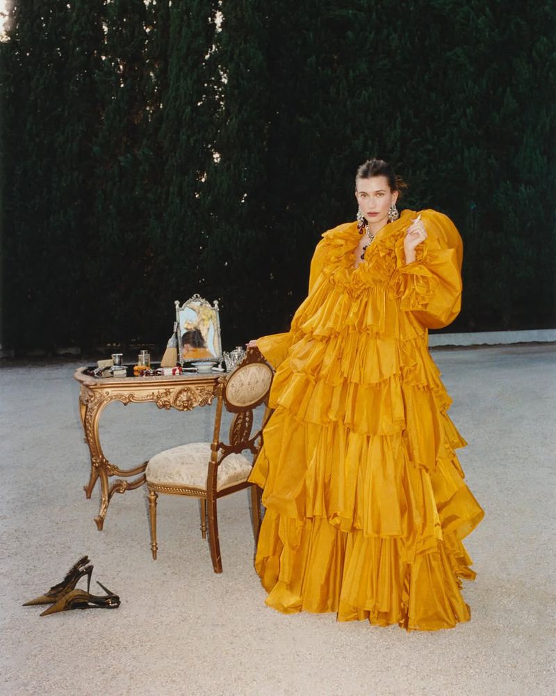Hailey Bieber wearing a voluminous ruffled yellow gown standing outdoors next to an ornate vanity table and mirror.