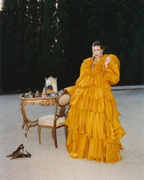 Hailey Bieber wearing a voluminous ruffled yellow gown standing outdoors next to an ornate vanity table and mirror.