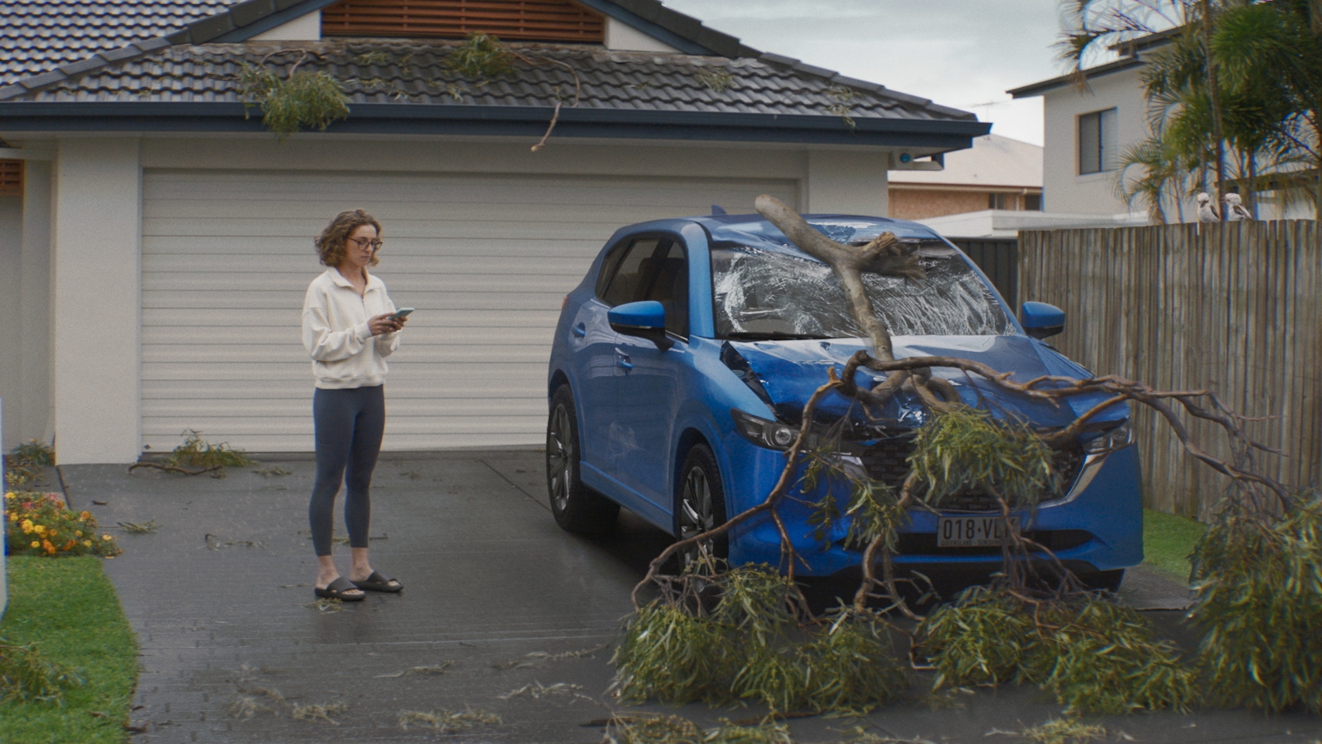 Woman on phone stands near damaged blue car covered in tree branches after a storm outside a home; house visible in background.