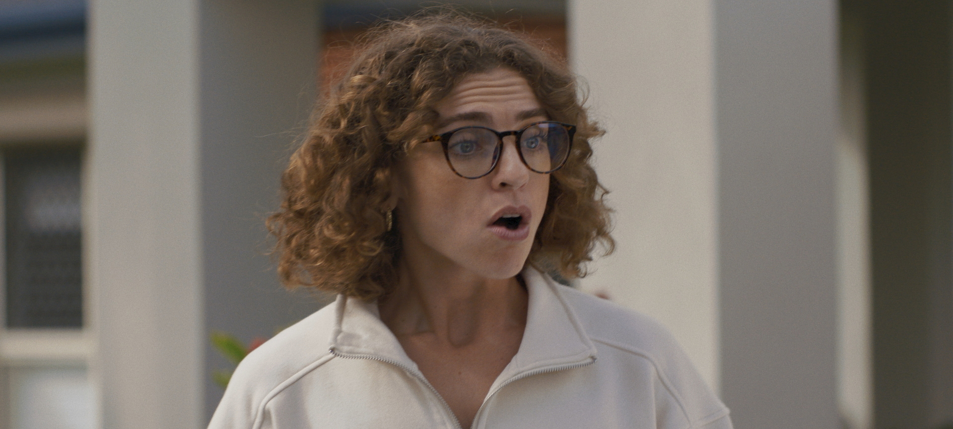 Close-up shot of a woman with curly hair and glasses wearing a light-colored sweater looking surprised, set against a neutral background.