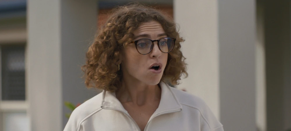 Close-up shot of a woman with curly hair and glasses wearing a light-colored sweater looking surprised, set against a neutral background.