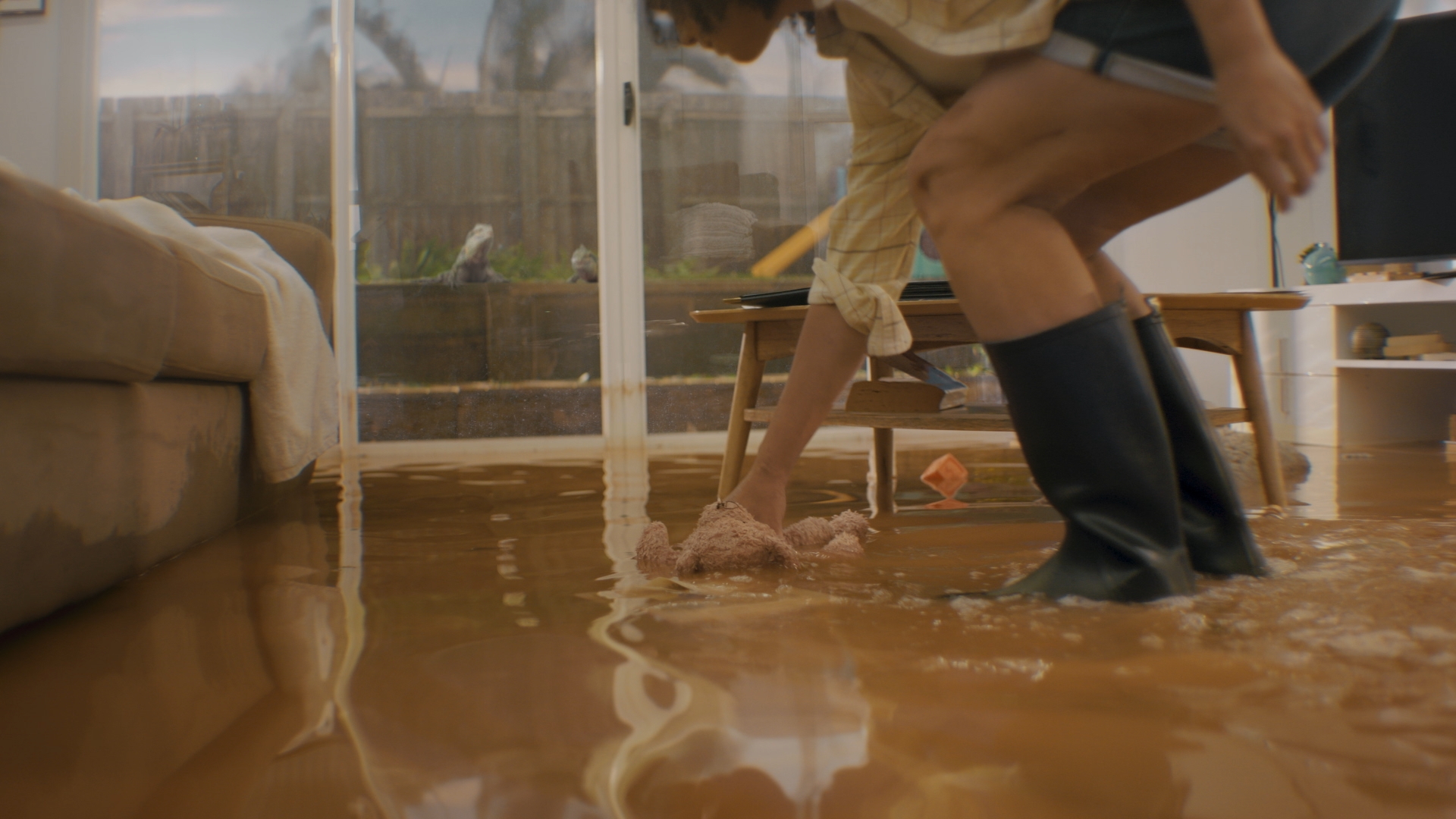 Adult wading through flooded living room wearing black rain boots, lifting up a tan stuffed animal out of the brown flood water.