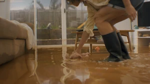 Person wearing black rain boots bending down in a living room flooded with muddy water, leaning over to pick up a stuffed toy.