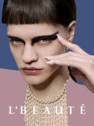 Close-up portrait of a model with dark blunt bangs, heavy black eye makeup, pearl necklace, and pink garment against a blue background