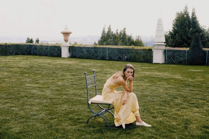 Model Grace Elizabeth in a pale yellow strapless dress sits on a metal chair in an expansive garden lawn.