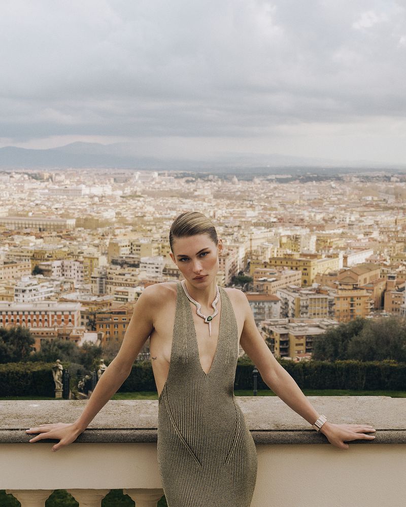 Model Grace Elizabeth in a plunging knit halter dress and statement necklace posing on a balcony overlooking an extensive cityscape.