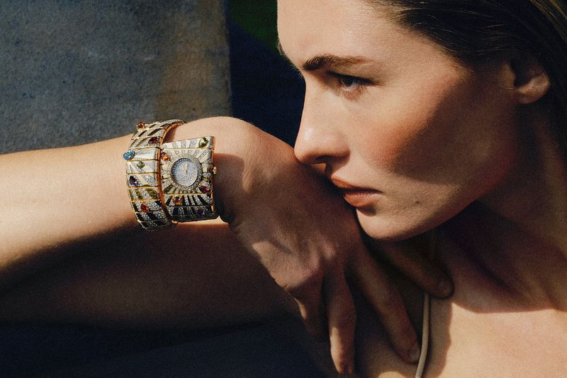 Model Grace Elizabeth rests her head on her arm wearing an ornate gold and diamond gemstone watch against a textured stone wall.