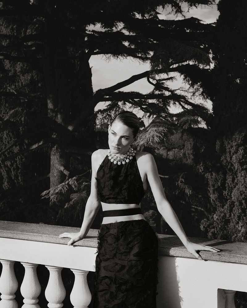 Grace Elizabeth wearing a textured black cutout dress and statement pearl collar necklace on a balcony overlooking trees.