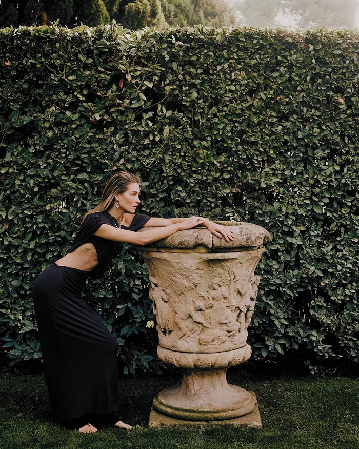 Grace Elizabeth wearing a black cut-out dress leaning on an ornate stone garden urn against a tall green hedge.