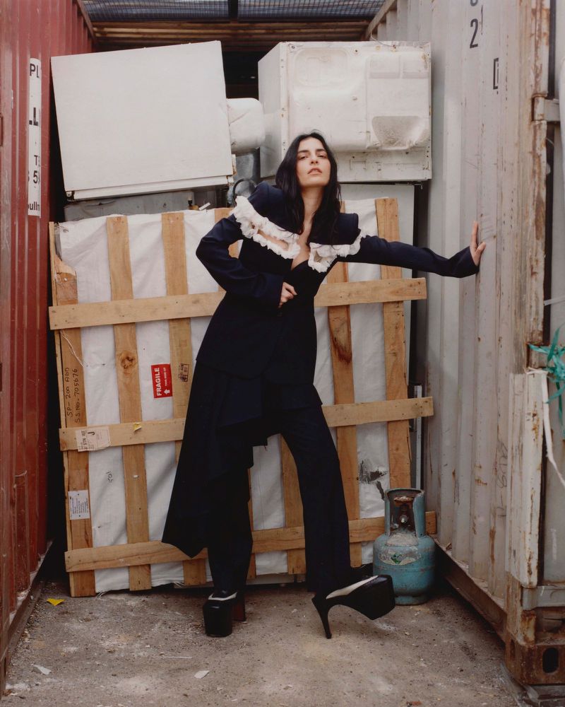 A stylish woman in a black outfit poses confidently in an industrial setting between shipping containers with stacked crates and tanks behind her.