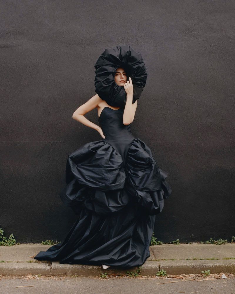 Woman in a large black dress and extravagant headpiece posing against a dark wall on a city street, fashion editorial shot.