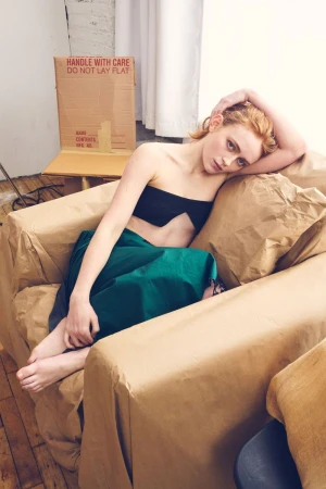Sadie Sink reclining barefoot in a kraft paper wrapped armchair wearing a black strapless crop top and emerald green skirt.
