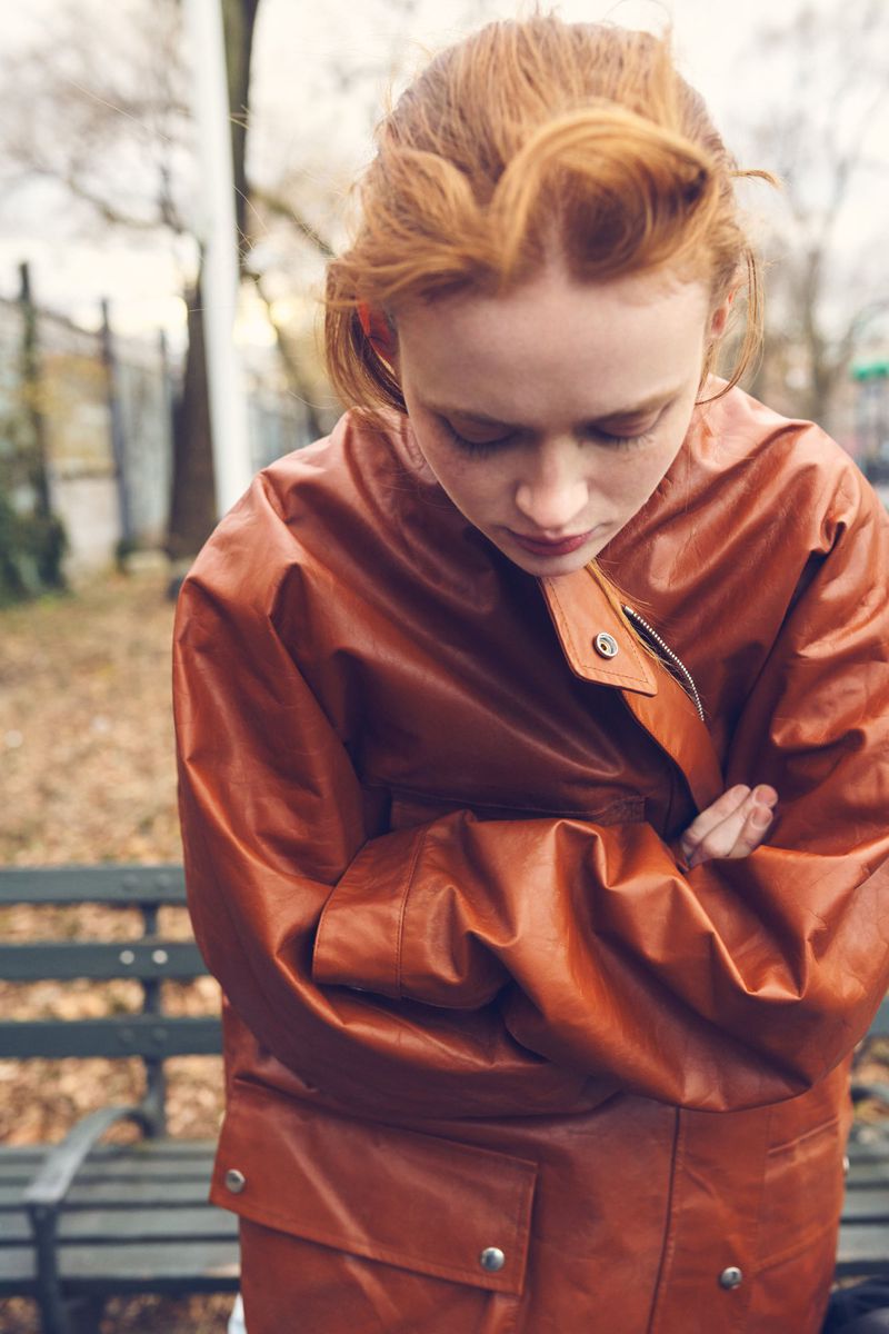 Sadie Sink looking down with arms crossed, wearing an oversized rust brown leather jacket outdoors in front of a park bench.
