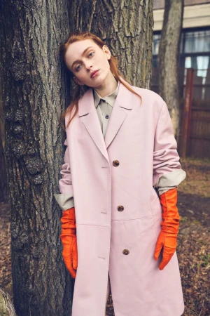 Sadie Sink wearing an oversized pastel pink coat and bright orange leather gloves while leaning against a textured tree trunk outdoors.