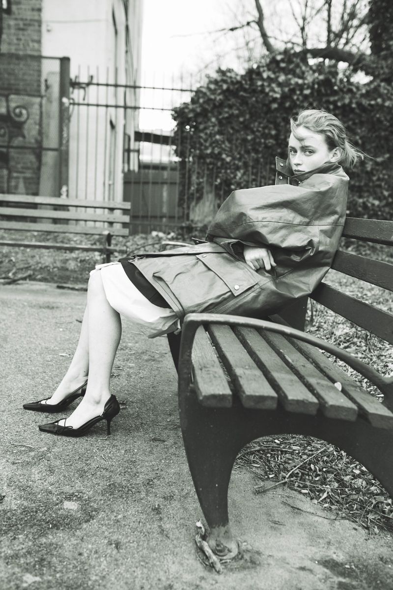Sadie Sink sits on a wooden park bench wearing an oversized utility jacket and pointed heels in a black and white portrait.