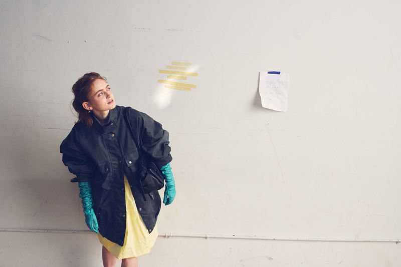 Sadie Sink in an oversized dark jacket, pale yellow dress, and long turquoise gloves posing against an off-white wall.