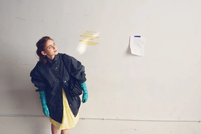 Sadie Sink in an oversized dark jacket, pale yellow dress, and long turquoise gloves posing against an off-white wall.