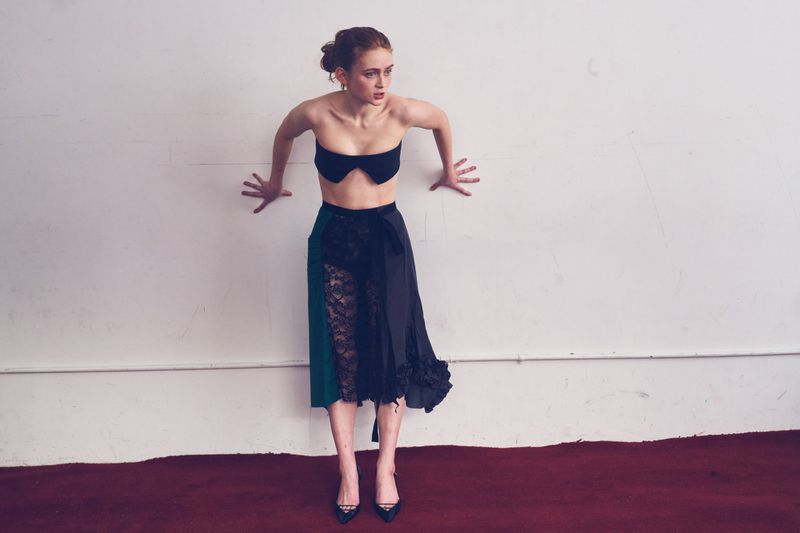 Sadie Sink poses against a white wall in a strapless black top, asymmetrical lace skirt, and black heels on a red carpet.