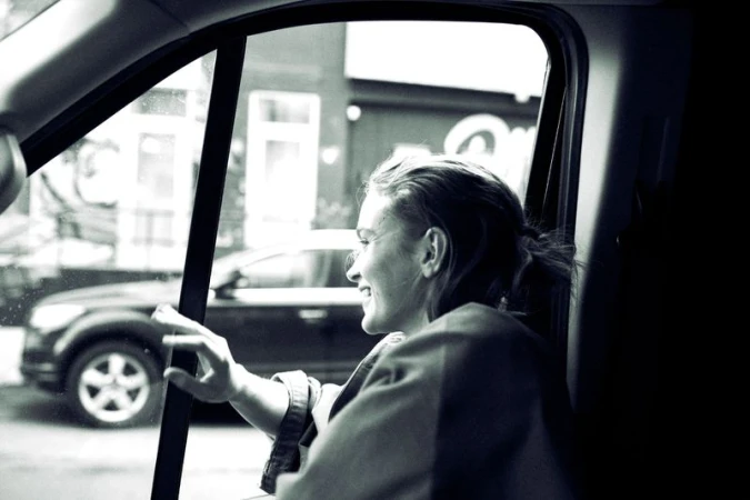 Actor Sadie Sink smiling as she looks out of an open car window in a black and white editorial shot.