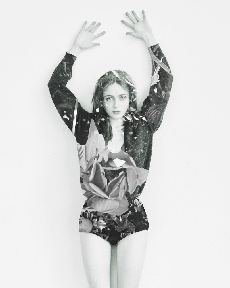 Sadie Sink posing in a floral patterned bodysuit with arms raised against a white studio background.