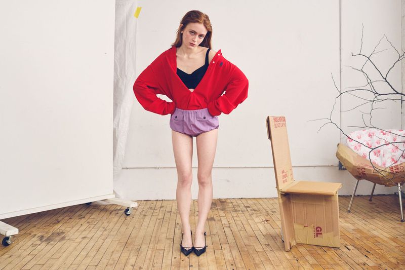 Sadie Sink poses wearing an open red jacket, black bra, lilac shorts, and black heels on a wooden floor in a studio.