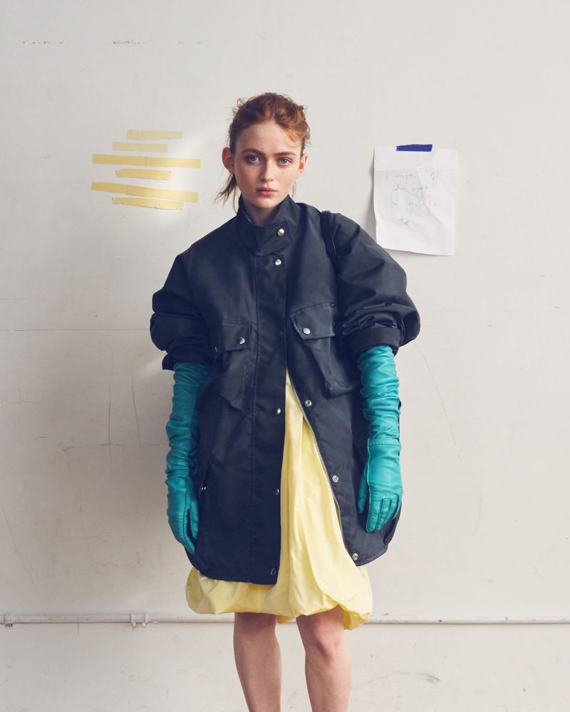 Sadie Sink wearing an oversized black jacket, pastel yellow dress, and long teal leather gloves against a studio wall.