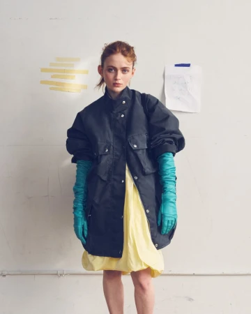 Sadie Sink wearing an oversized black jacket, pastel yellow dress, and long teal leather gloves against a studio wall.