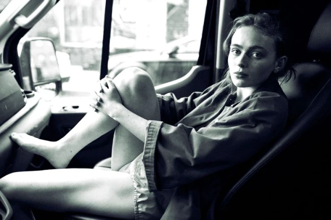 Sadie Sink sitting barefoot in the passenger seat of a vehicle with knees drawn up, wearing an oversized buttoned jacket.