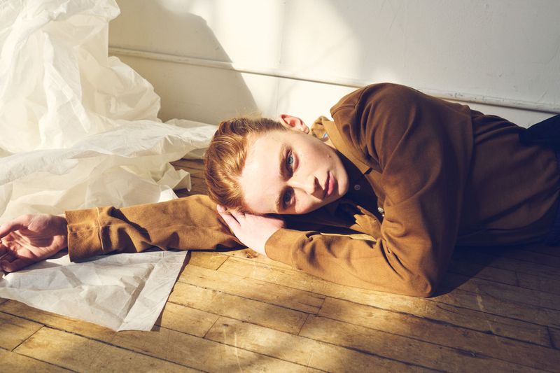 Sadie Sink lying on a wooden floor wearing a brown jacket next to crumpled white paper in sunlight.