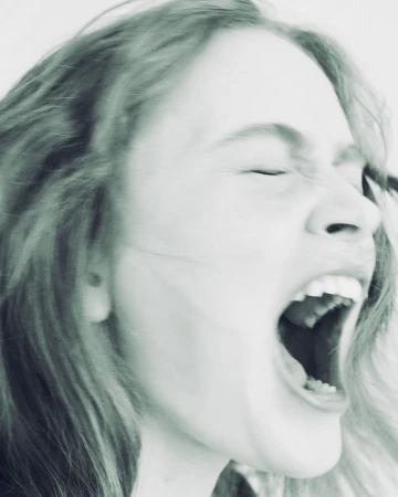 Close-up portrait of actor Sadie Sink screaming with mouth wide open against a pale background.