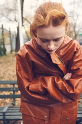 Sadie Sink looking down with arms crossed, wearing an oversized rust brown leather jacket outdoors in front of a park bench.