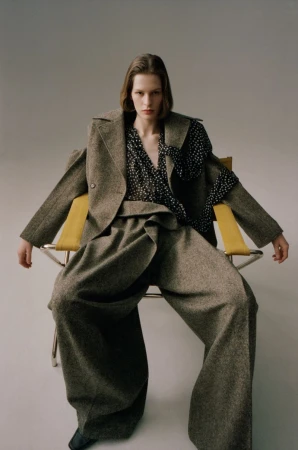 Model Hannah Weber seated in an oversized tweed suit and polka dot blouse on a yellow-accented chair against a studio backdrop.