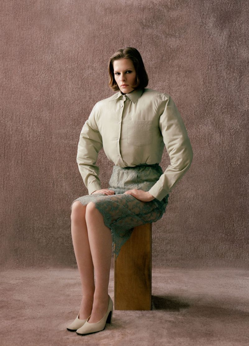 Model Hannah Weber seated on a wooden block wearing a padded beige button-up shirt and green lace pencil skirt against a textured backdrop.
