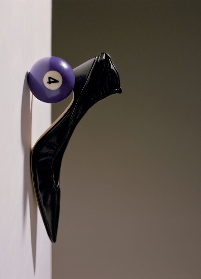 A glossy black pointed-toe high heel balanced vertically against a purple number four pool ball along a wall edge.
