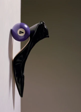 A glossy black pointed-toe high heel balanced vertically against a purple number four pool ball along a wall edge.