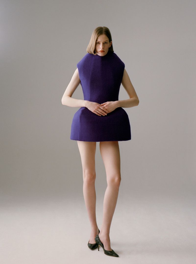 Model Hannah Weber in a sculptural purple geometric mini dress and dark pointed heels against a studio backdrop.