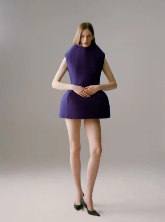 Model Hannah Weber in a sculptural purple geometric mini dress and dark pointed heels against a studio backdrop.