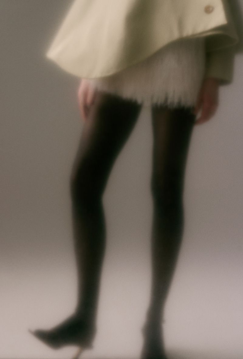 Cropped view of a model in black tights, pointed heels, and a pale structured coat in a soft-focus studio setting.