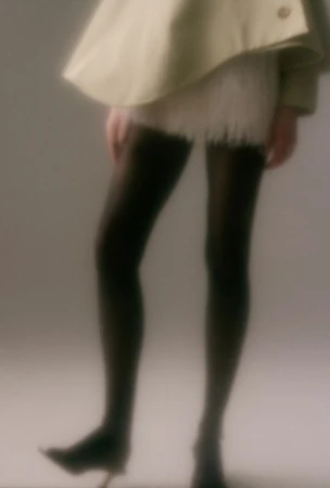 Cropped view of a model in black tights, pointed heels, and a pale structured coat in a soft-focus studio setting.