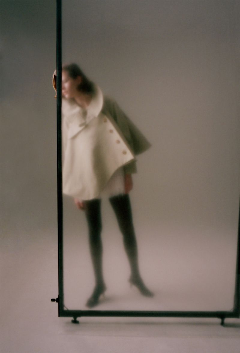 Model wearing a cream cape jacket and dark tights standing behind a translucent studio screen.
