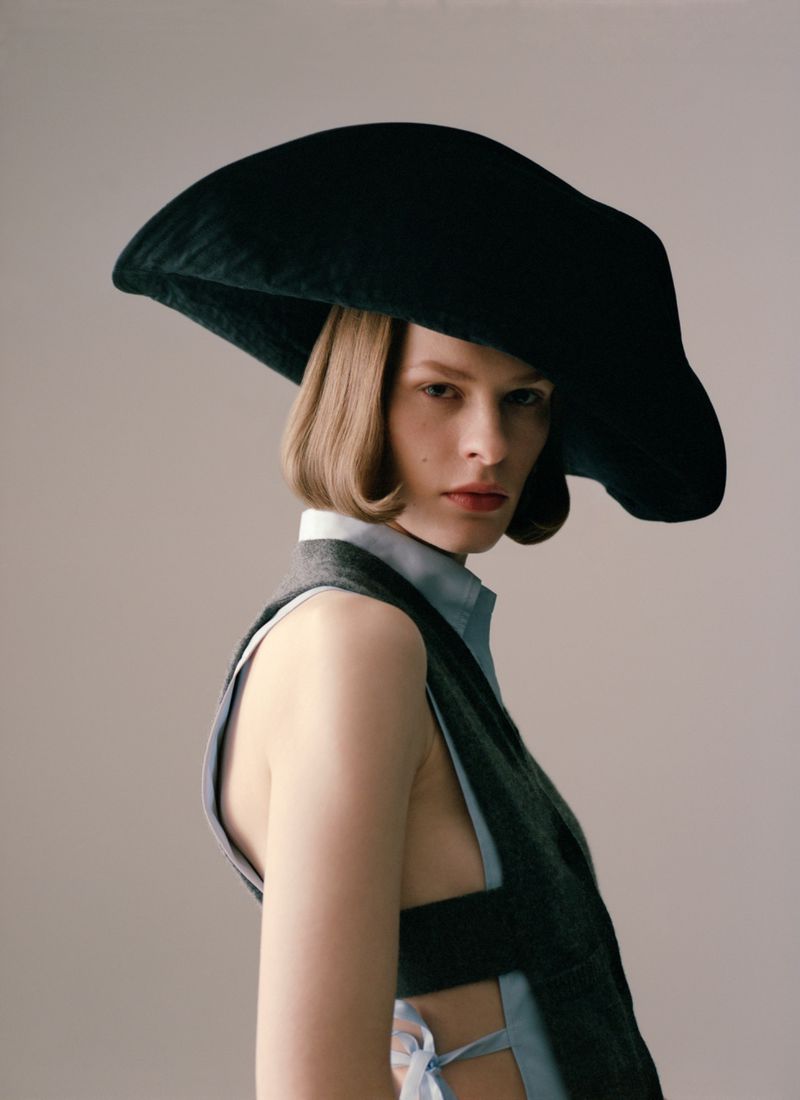 Model wearing a large sculptural dark hat and an open-sided vest against a plain grey studio backdrop.