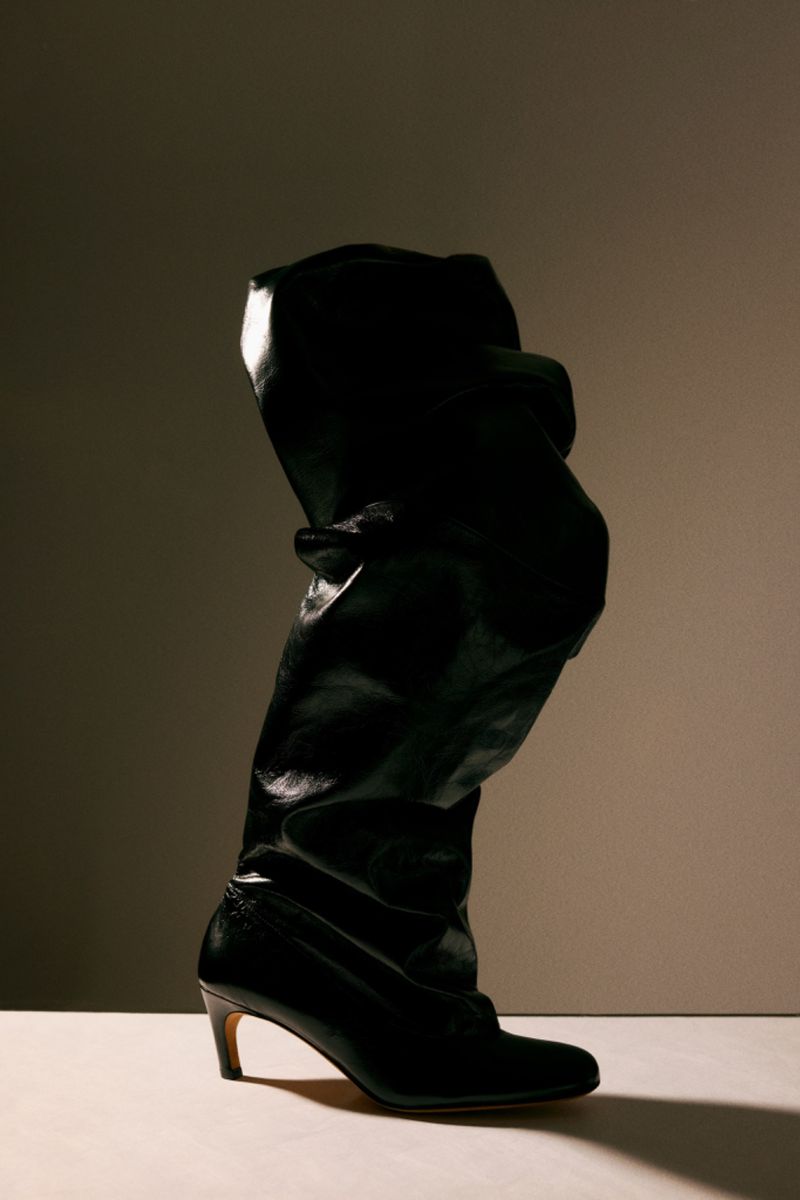 Slouchy black leather knee-high boot with a kitten heel standing upright under dramatic studio lighting against a gradient backdrop.